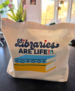 Cream canvas tote bag with green handles featuring a ‘Libraries Are Life’ graphic: a stylized stack of books in blue, yellow, and green beneath the word ‘Libraries’ in script and ‘ARE LIFE’ in block letters.