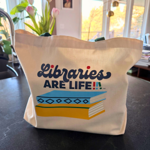 “Cream canvas tote bag with green handles featuring a ‘Libraries Are Life’ graphic: a stylized stack of books in blue, yellow, and green beneath the word ‘Libraries’ in script and ‘ARE LIFE’ in block letters.”