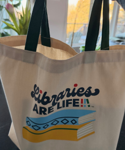 Cream canvas tote bag with green handles featuring a ‘Libraries Are Life’ graphic: a stylized stack of books in blue, yellow, and green beneath the word ‘Libraries’ in script and ‘ARE LIFE’ in block letters.
