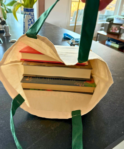 Open cream canvas tote bag with green handles sitting on a kitchen counter, filled with several stacked books.
