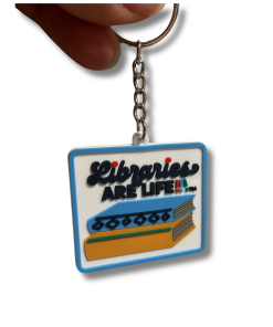 Close-up of a rectangular Libraries Are Life keychain featuring a white background, blue border, and raised lettering above a colorful stack of books. The design includes black text reading “Libraries Are Life” with small multicolored accents. A silver metal chain and split key ring are attached at the top.