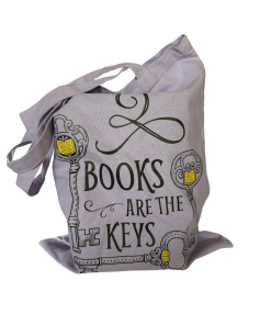 Lavender canvas tote bag with long shoulder straps, filled with books and featuring the phrase “Books Are the Keys” in black decorative lettering, surrounded by ornate skeleton key illustrations with yellow book accents, against a white background.