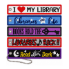 Five rectangular embroidered fabric keychain patches arranged vertically on a white background, each with textured stitching, colorful borders, and a metal grommet. Designs include “I Love My Library” in red and white, “Libraries Are Life” in blue, “Books Hold the Key” with a skeleton key on purple, “Libraries Rock!!” in red and black, and “Read After Dark” with a crescent moon and stars on black and purple. A silver ball chain is attached to the bottom patch.