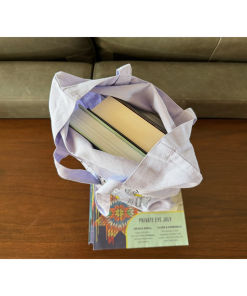 Overhead view of an open lavender canvas tote bag filled with several hardcover books, demonstrating its storage capacity. The bag rests on a stack of reading materials on a wooden table, with a green sofa in the background.