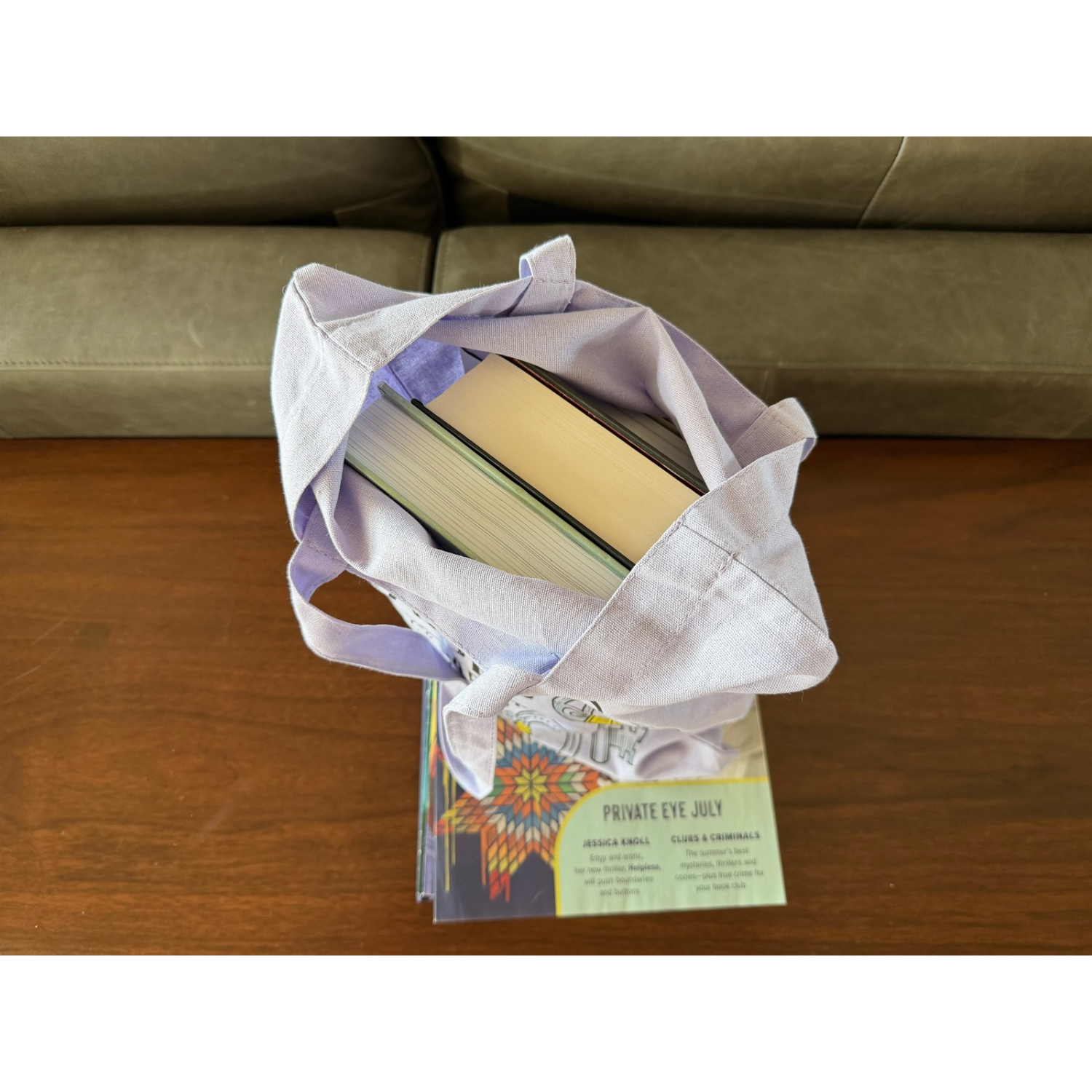 Overhead view of an open lavender canvas tote bag filled with several hardcover books, demonstrating its storage capacity. The bag rests on a stack of reading materials on a wooden table, with a green sofa in the background.