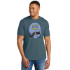 Model wearing a slate blue unisex crewneck T-shirt featuring Laurie Conley’s 2027 “Mysteries Await at Your Library” artwork. The design shows Inspector Whiskers, a black detective cat wearing a yellow hat and scarf, holding a magnifying glass beside a library book return slot. Yellow and black lettering appears against a lavender background. Model shown wearing blue jeans against a white background.