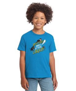 Smiling child modeling a bright blue youth crewneck T-shirt featuring the 2027 “Mysteries Await at Your Library” slogan in yellow and white lettering, surrounded by Laurie Conley’s playful mystery-themed characters, including a detective cat, friendly ghost, and other mysterious figures. Short-sleeved shirt shown with light blue jeans against a white background.