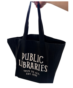 Black canvas tote bag with long shoulder straps, held up by one hand to show its size and shape. Cream-colored vintage-style lettering reads “PUBLIC LIBRARIES,” with “OPEN TO ALL” and “EST. 1732” beneath.