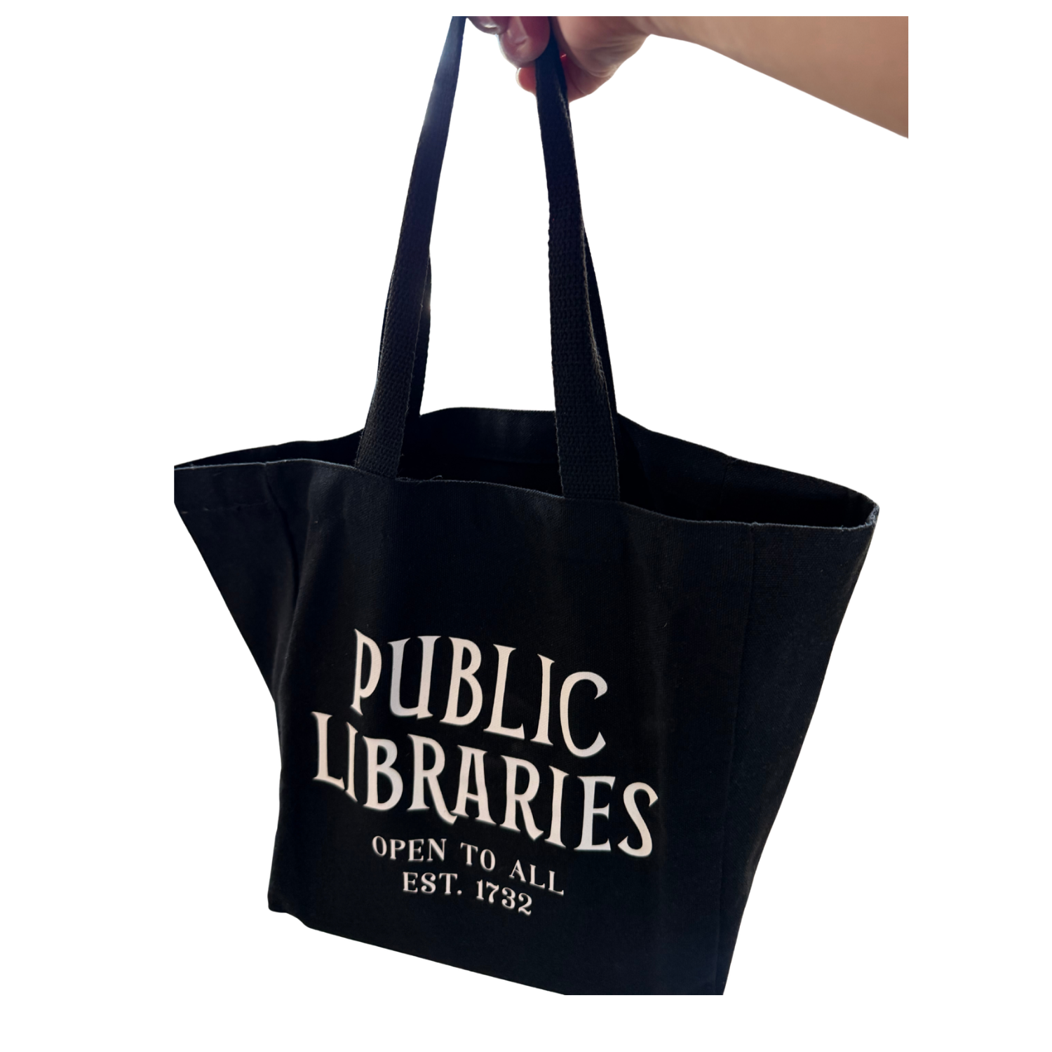 Black canvas tote bag with long shoulder straps, held up by one hand to show its size and shape. Cream-colored vintage-style lettering reads “PUBLIC LIBRARIES,” with “OPEN TO ALL” and “EST. 1732” beneath.