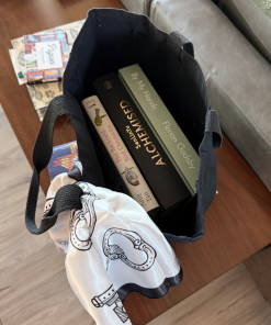Overhead view of an open black canvas tote bag holding three hardcover books, including The Backyard Bird Chronicles, Alchemy, and By My Hands. A white scarf with black illustrated designs is draped over one handle, demonstrating the bag's roomy interior and capacity for carrying books.