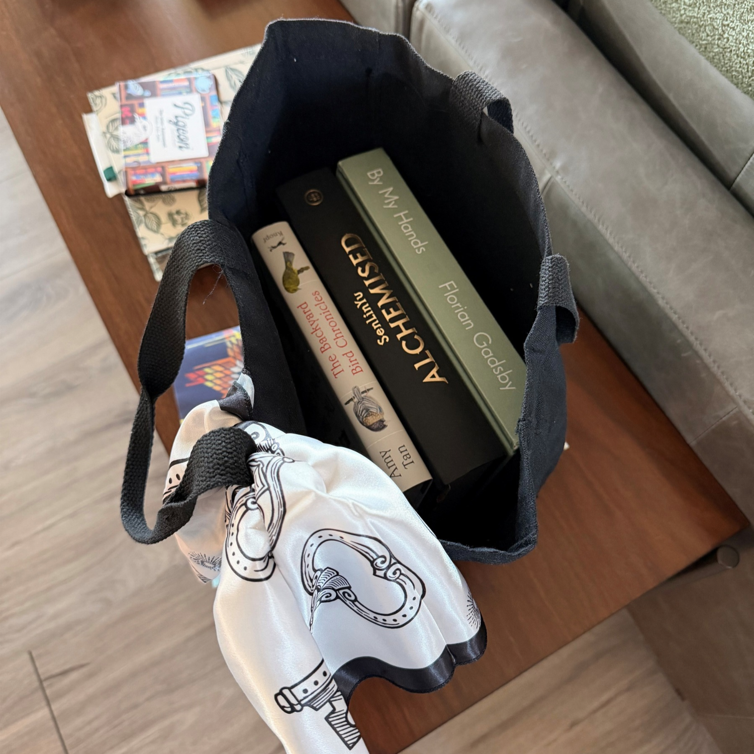 Overhead view of an open black canvas tote bag holding three hardcover books, including The Backyard Bird Chronicles, Alchemy, and By My Hands. A white scarf with black illustrated designs is draped over one handle, demonstrating the bag's roomy interior and capacity for carrying books.