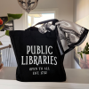 Black canvas tote bag displayed on a countertop, featuring vintage-style white lettering reading “PUBLIC LIBRARIES – OPEN TO ALL – EST. 1732.” A white satin scarf with black illustrated skeleton keys is tied around one handle, adding a decorative touch.