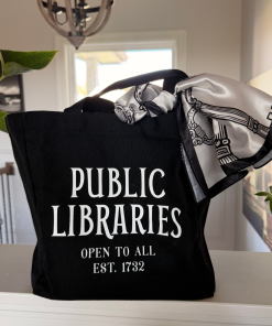 Black canvas tote bag displayed on a countertop, featuring vintage-style white lettering reading “PUBLIC LIBRARIES – OPEN TO ALL – EST. 1732.” A white satin scarf with black illustrated skeleton keys is tied around one handle, adding a decorative touch.