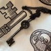 Black skeleton key necklace with a round, four-hole decorative bow and matching black chain. The key features the white inscription “Books Hold the Key to My Heart” and rests on a cream-colored scarf with black illustrated key and book designs.