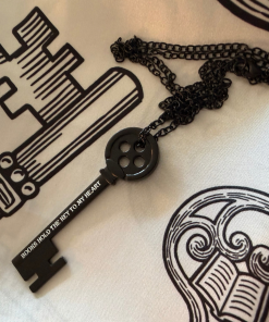 Black skeleton key necklace with a round, four-hole decorative bow and matching black chain. The key features the white inscription “Books Hold the Key to My Heart” and rests on a cream-colored scarf with black illustrated key and book designs.