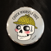 Round silver compact mirror featuring Laurie Conley’s illustration of a smiling skull with rosy cheeks wearing a yellow plaid detective hat. The words “Check Yourself Out!” arch above the skull, with “CSLP 2027” printed below. The reflective compact rests on a black fabric background.