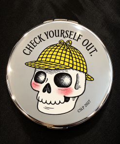 Round silver compact mirror featuring Laurie Conley’s illustration of a smiling skull with rosy cheeks wearing a yellow plaid detective hat. The words “Check Yourself Out!” arch above the skull, with “CSLP 2027” printed below. The reflective compact rests on a black fabric background.