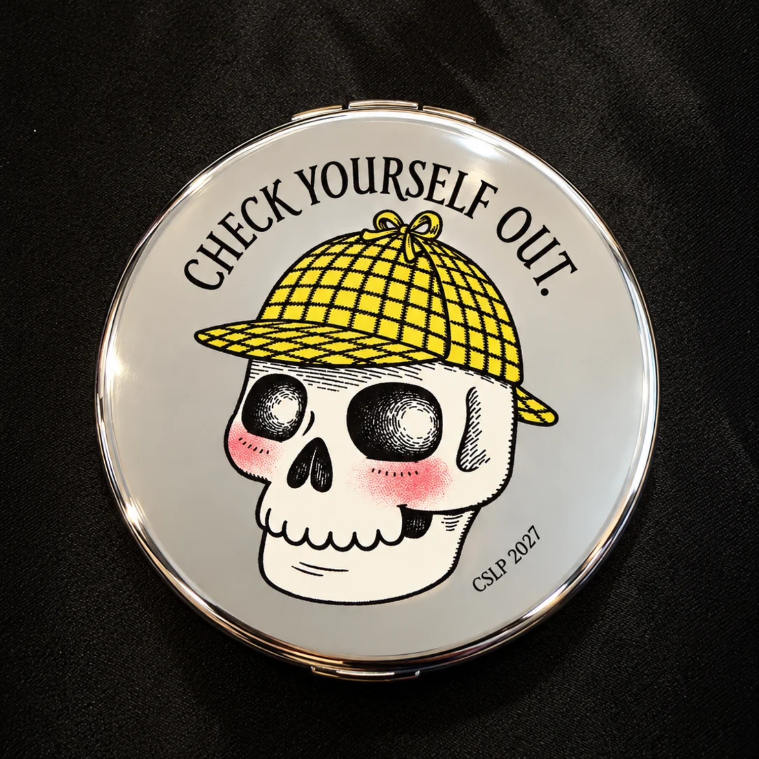 Round silver compact mirror featuring Laurie Conley’s illustration of a smiling skull with rosy cheeks wearing a yellow plaid detective hat. The words “Check Yourself Out!” arch above the skull, with “CSLP 2027” printed below. The reflective compact rests on a black fabric background.
