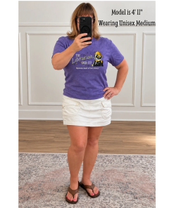 Woman modeling a heather purple unisex crewneck T-shirt featuring Laurie Conley’s “The Librarian Did It!” design with Inspector Whiskers, a black detective cat wearing a yellow deerstalker hat and holding a magnifying glass. The “Mysteries Await at Your Library” slogan appears beneath the artwork. Paired with a white cargo-style skirt and brown sandals, shown in a full-length mirror selfie against a neutral indoor background.