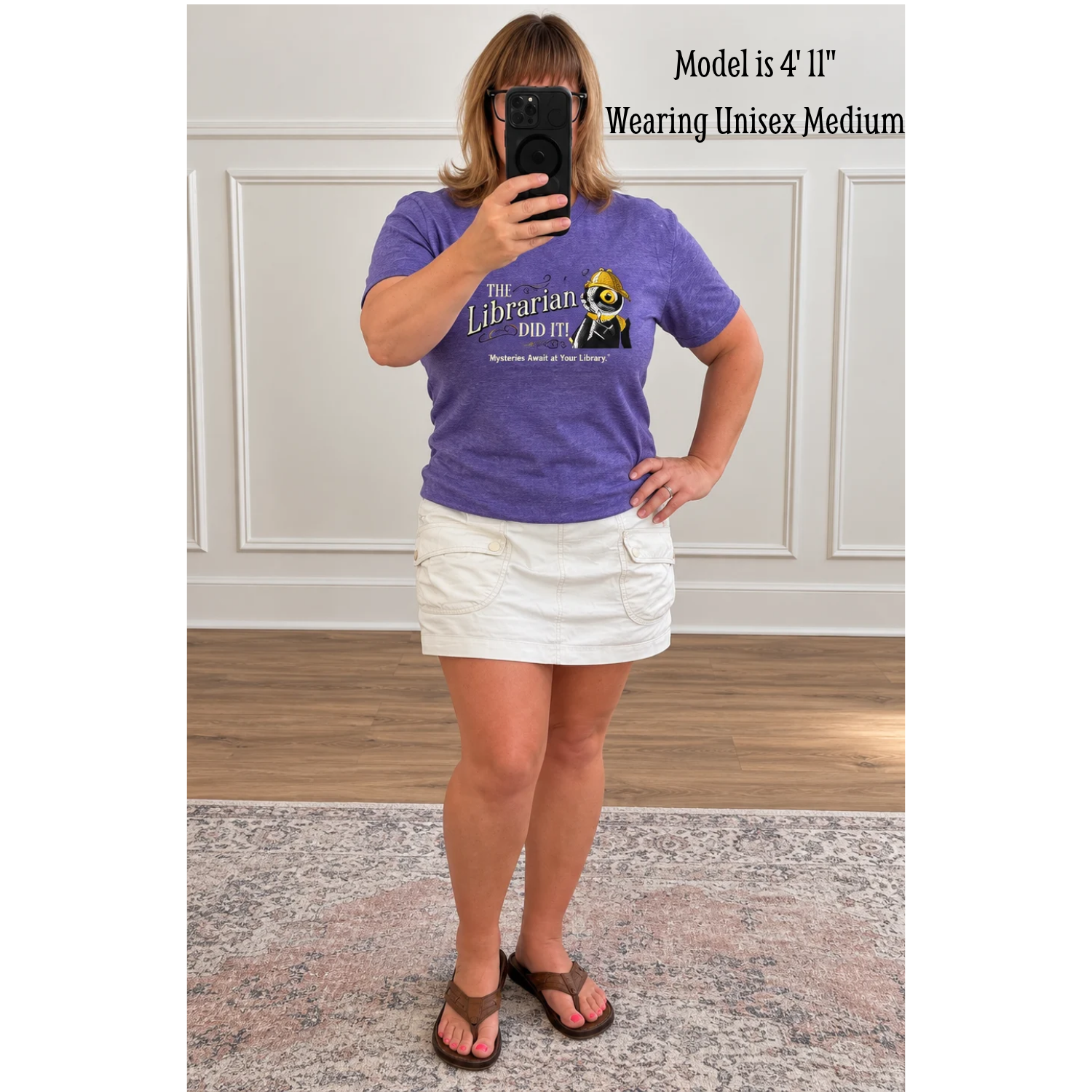 Woman modeling a heather purple unisex crewneck T-shirt featuring Laurie Conley’s “The Librarian Did It!” design with Inspector Whiskers, a black detective cat wearing a yellow deerstalker hat and holding a magnifying glass. The “Mysteries Await at Your Library” slogan appears beneath the artwork. Paired with a white cargo-style skirt and brown sandals, shown in a full-length mirror selfie against a neutral indoor background.
